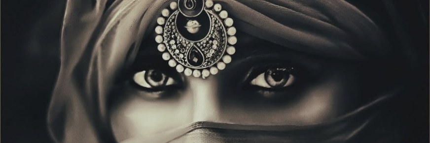 A veiled woman&rsquo;s eyes beneath an ornate maang tikka, in sepia